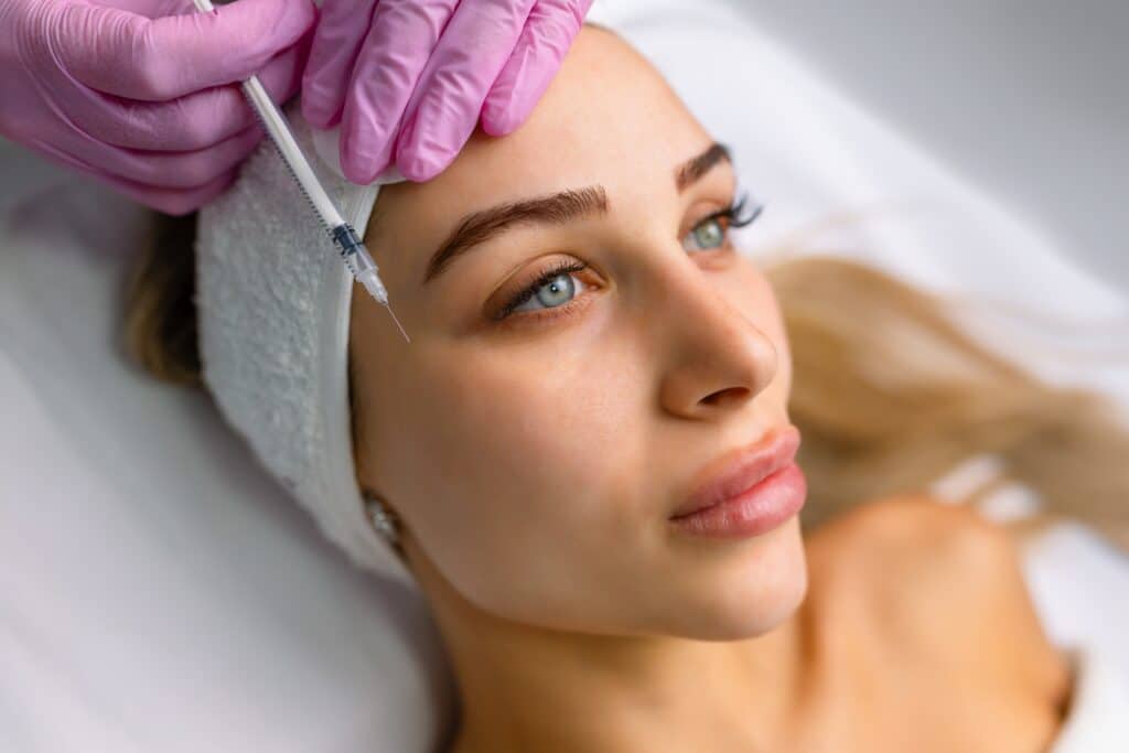 Beautiful woman, wearing a spa headband, looks straight ahead as her injector administers Botox and fillers